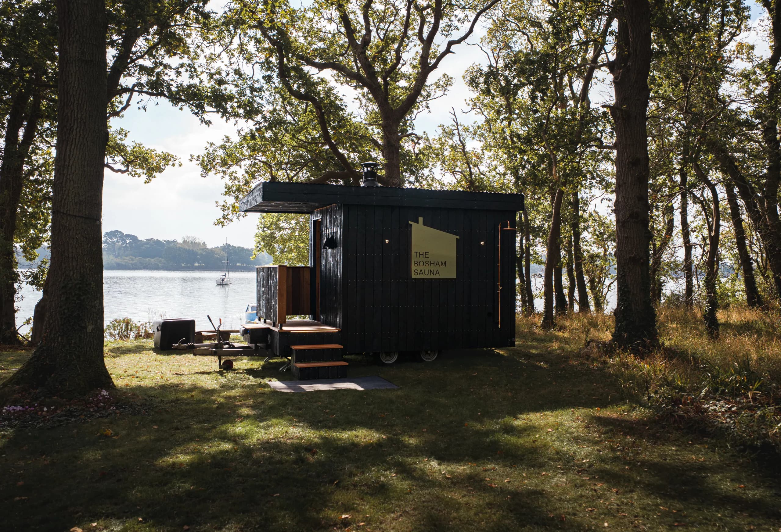 The black timber Bosham Sauna set up among oak trees, with the harbour behind.