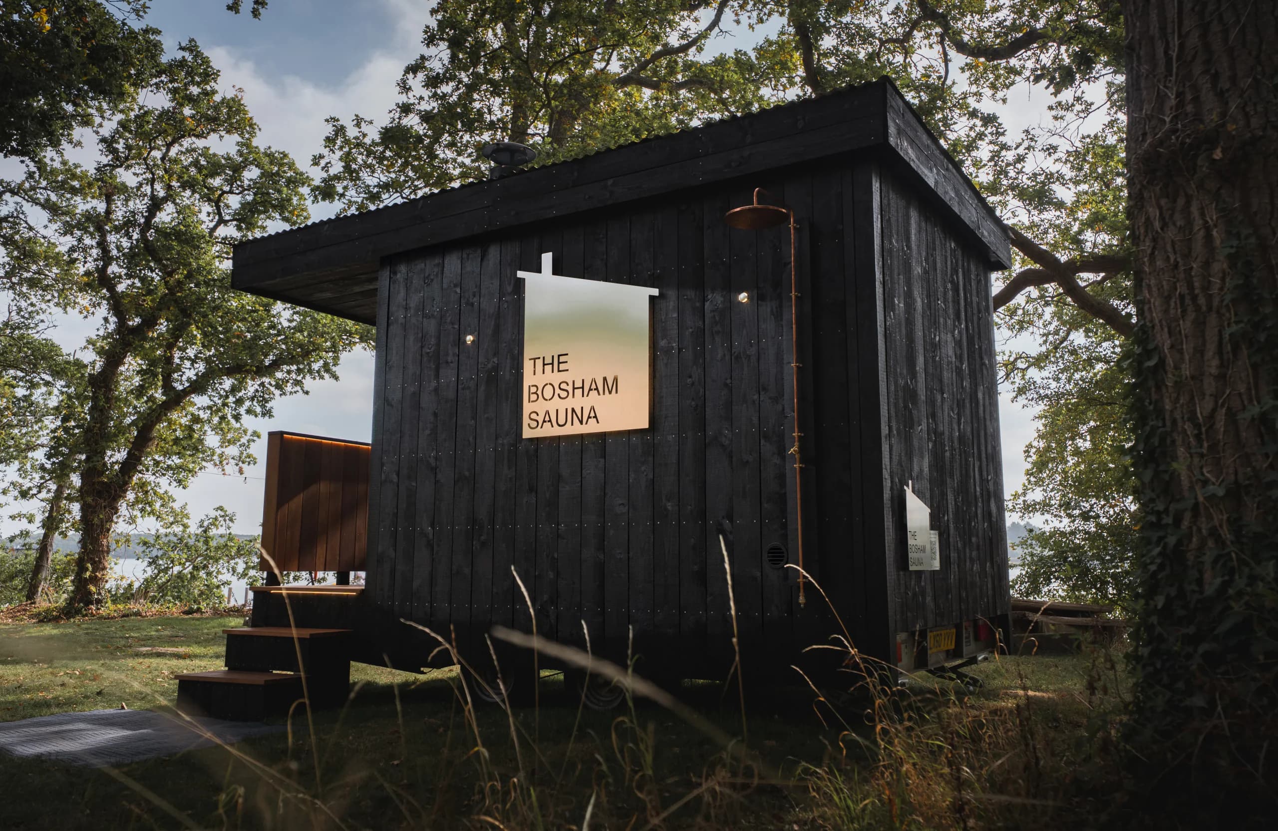 The Bosham Sauna under the oaks: black timber cladding, cedar steps, a copper shower and the house-shaped nameplate.